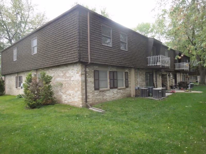 Two-story brick and wood-sided apartment building with small balconies, HVAC units, and a grassy lawn in front.