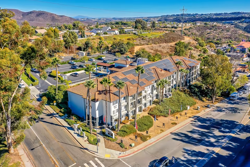 Aerial view of a senior living facility building with a red-tiled roof and solar panels, surrounded by palm trees and landscaping. The building is located at a street corner with visible parking lots and hills in the background under a clear blue sky.