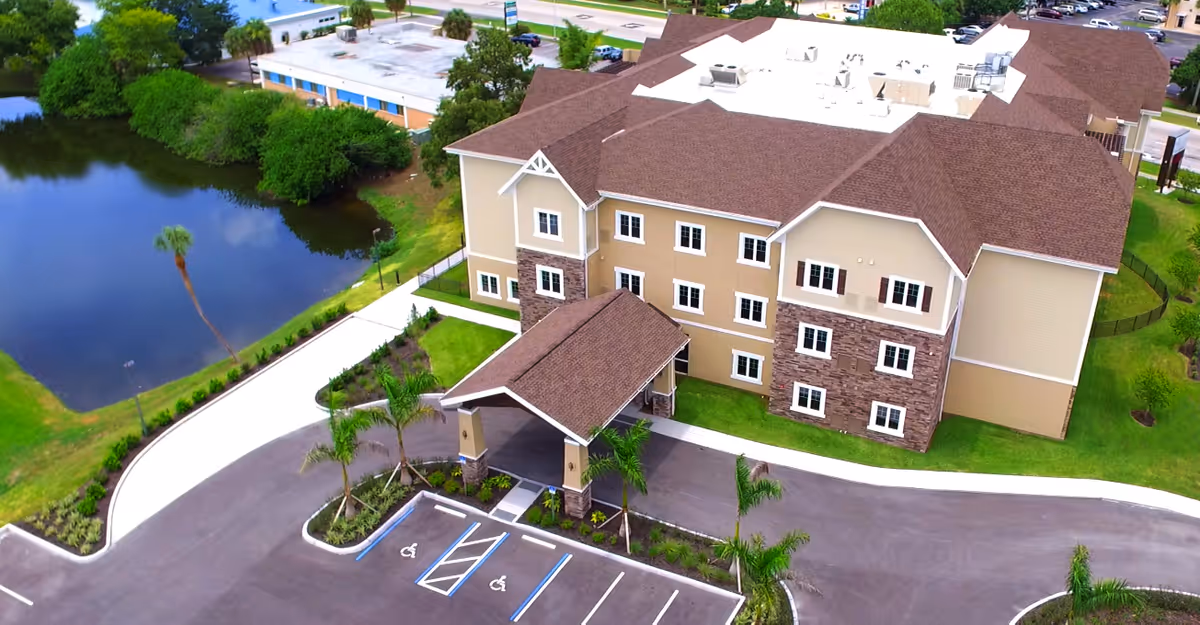 Aerial view of a three-story assisted living building with a covered entrance, parking spaces and a pond nearby.