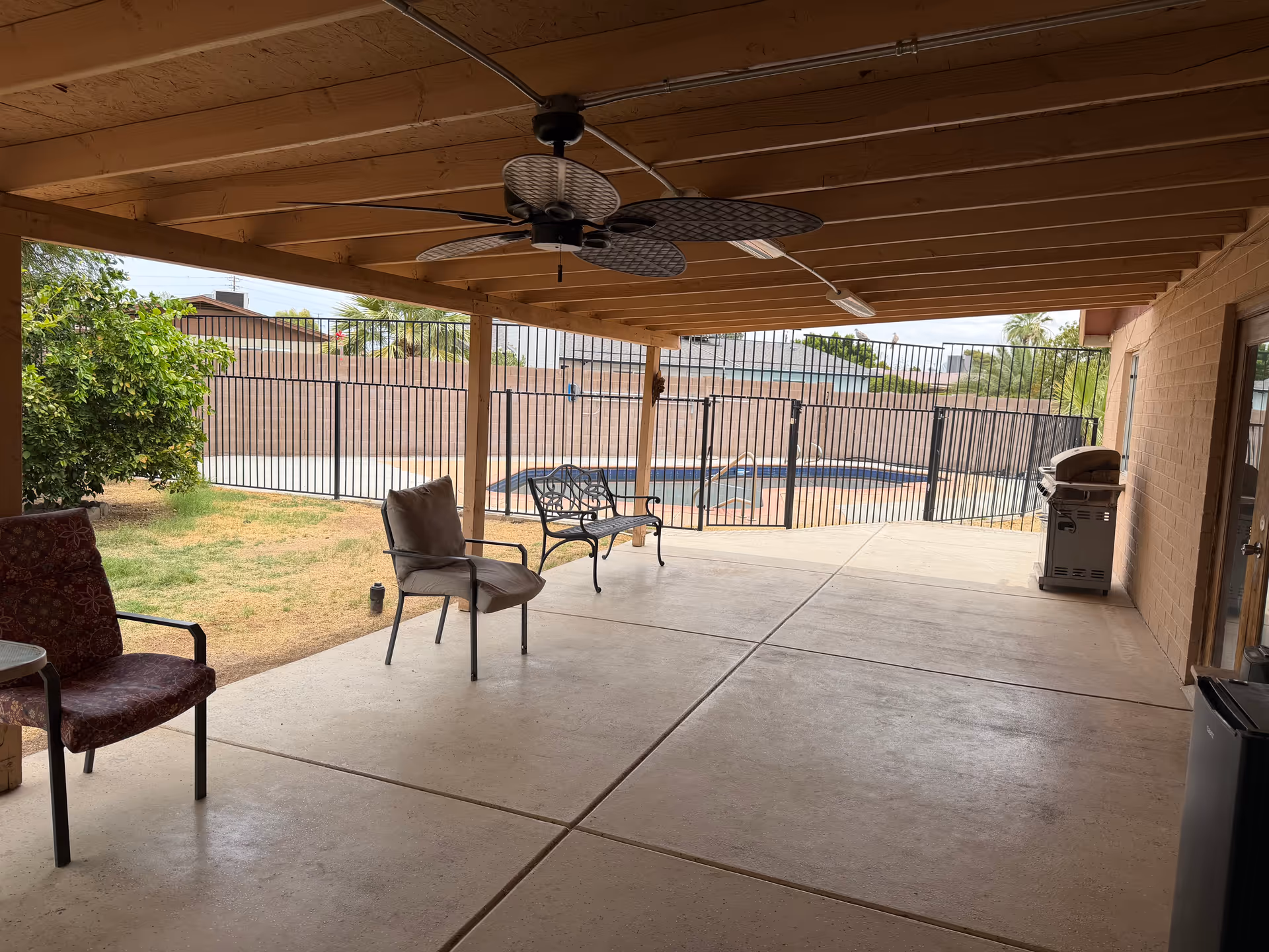 Covered patio with outdoor seating, ceiling fan, grill, and a fenced pool in background.