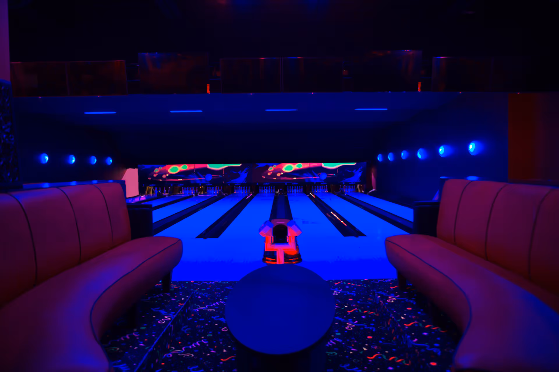Dim bowling alley with neon blue lighting, curved seating on both sides and carpeted floor.