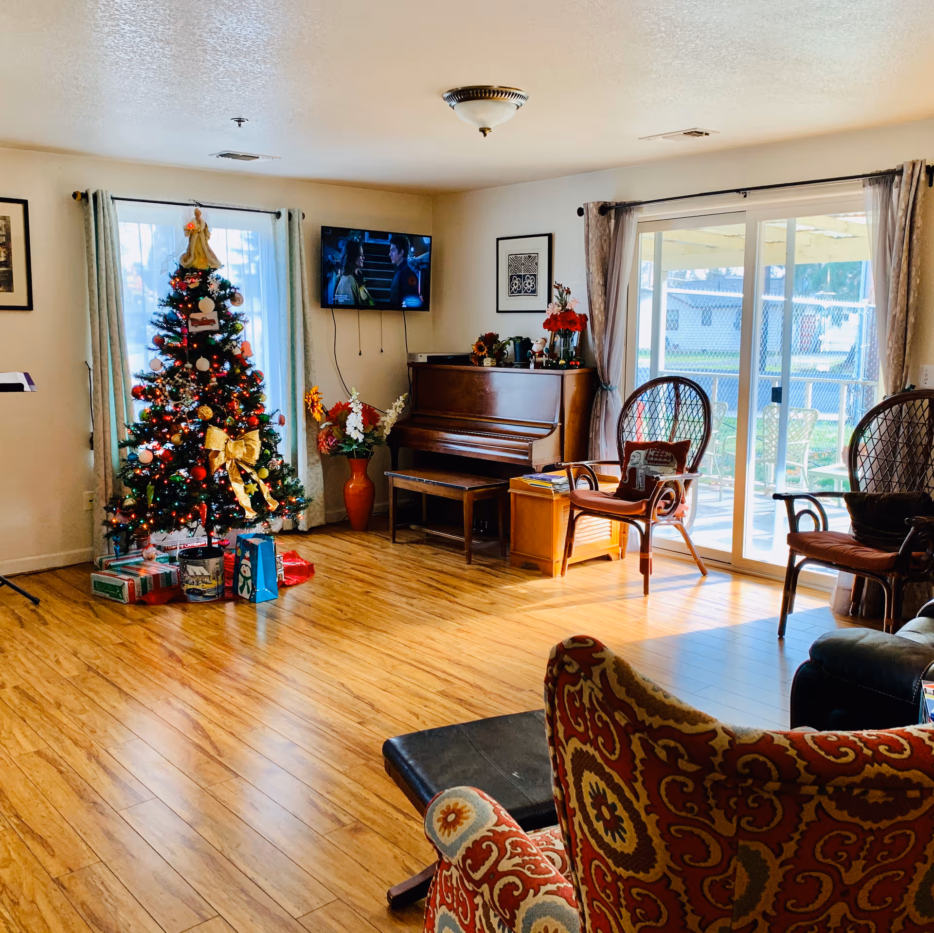 A cozy living room decorated for Christmas with a Christmas tree adorned with ornaments and lights, presents underneath, a wooden piano with a bench, two wicker chairs with cushions, a patterned armchair, and a flat-screen TV mounted on the wall. Large windows and sliding glass doors let in natural light and show an outdoor patio area.