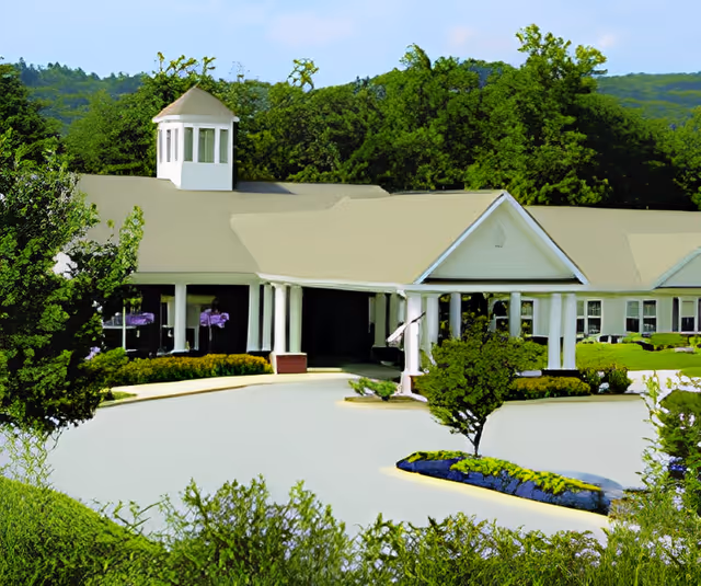Exterior view of a single-story building with a light-colored roof and white columns at the entrance, surrounded by greenery and trees under a clear blue sky.