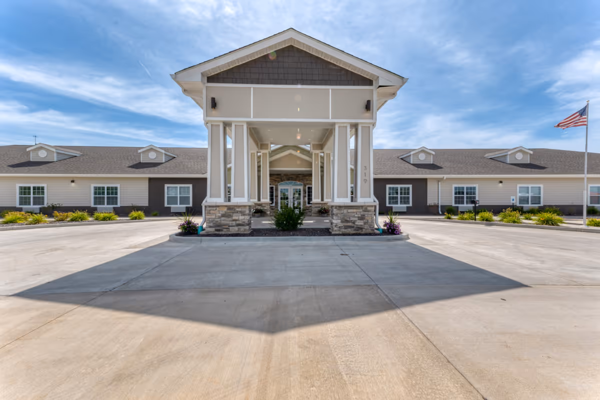 Front exterior view of High Point Residence Quincy assisted living facility in Illinois, featuring a covered entrance with stone pillars, a wide driveway, landscaped greenery, and an American flag on a flagpole to the right.
