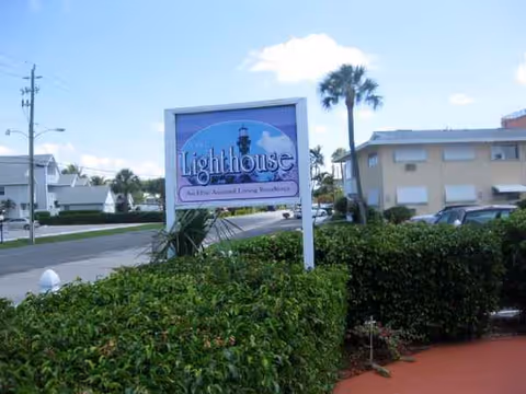 Exterior view of Belair by the Sea with a blue Lighthouse sign, palm trees, and shrubs.