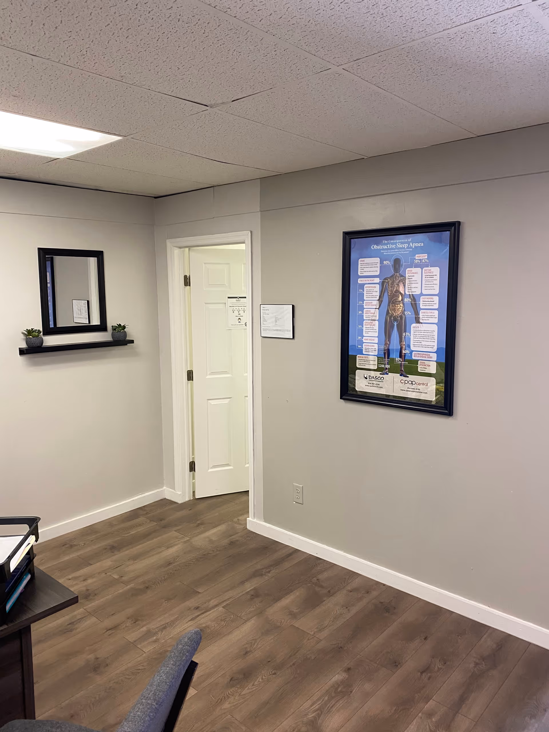 A small medical office corner with door, framed poster, mirror, shelf, and potted plants.