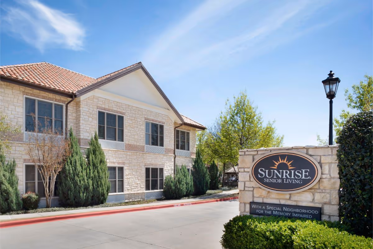 Exterior view of a two-story stone Sunrise Senior Living building with landscaping and a sign at the entrance.