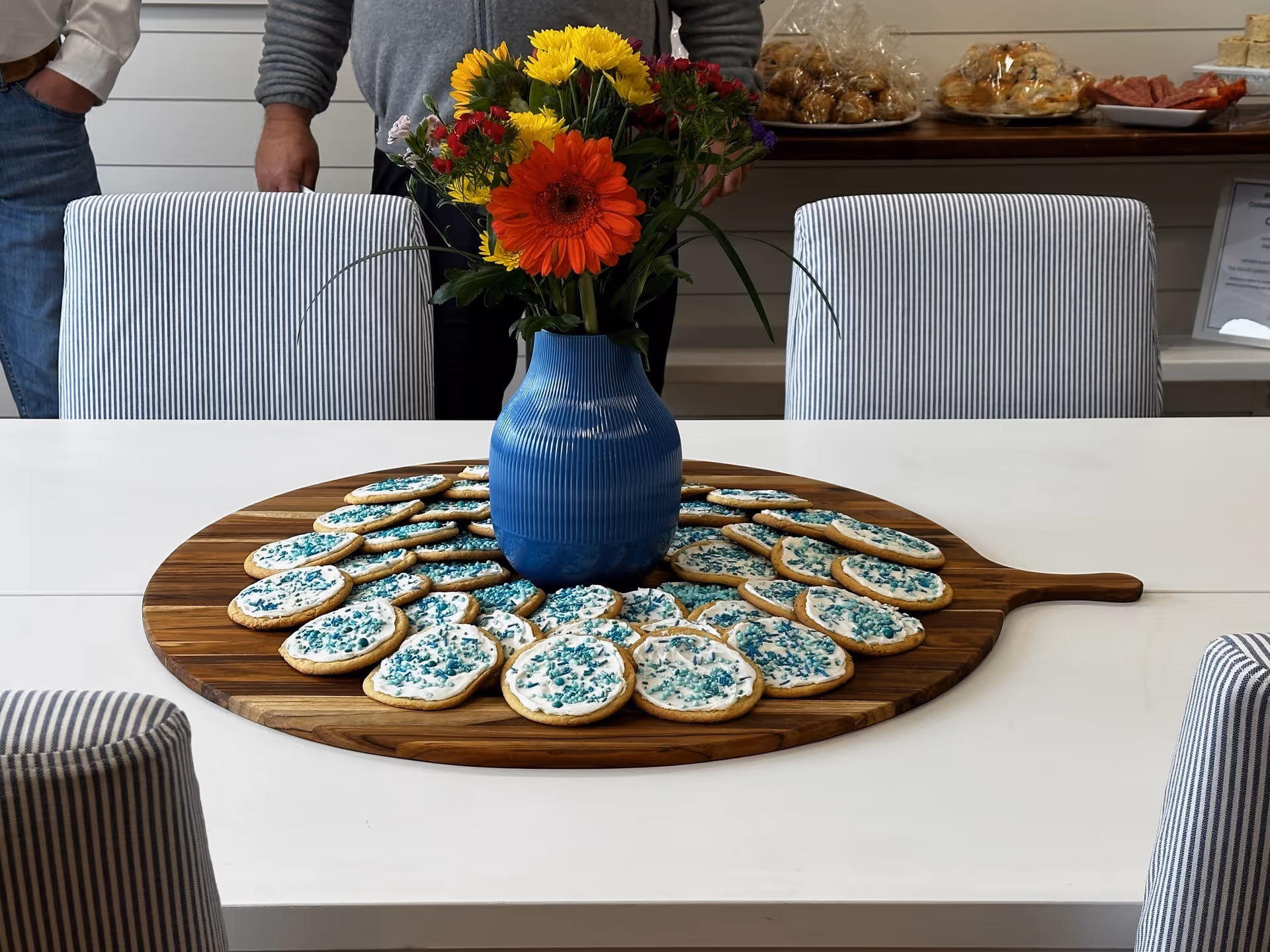 Blue vase with colorful flowers on a white table surrounded by cookies on a round board.