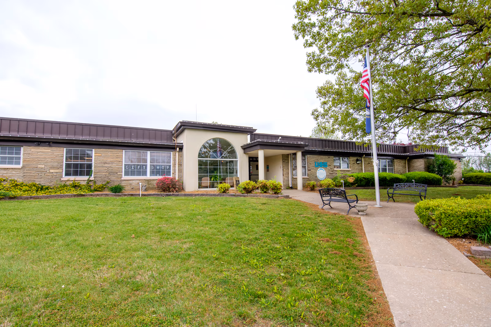 Front exterior of a single-story hospital-like building with lawn, flagpole, and benches.