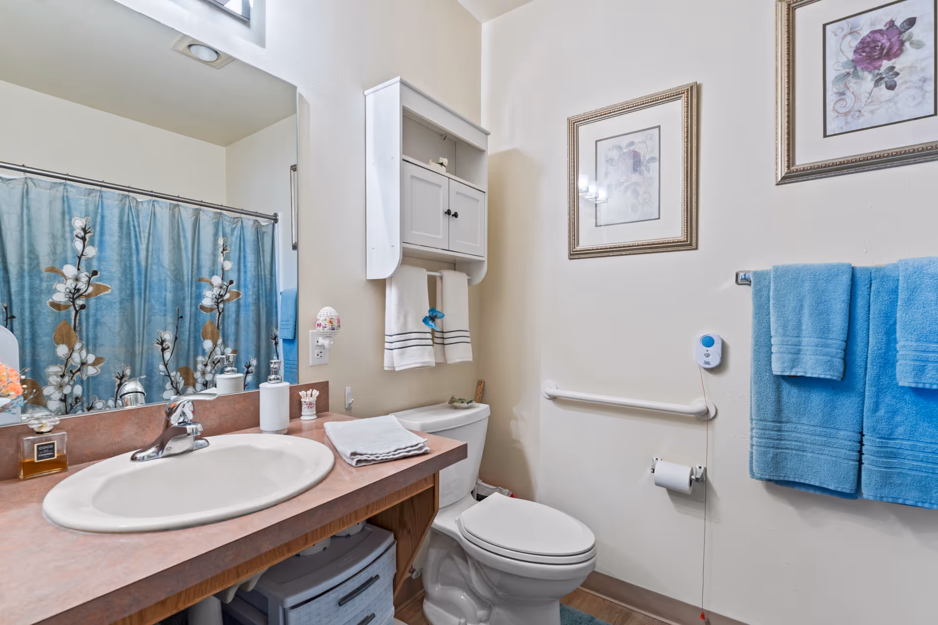 A clean and well-lit bathroom featuring a white sink with a silver faucet on a pinkish countertop, a toilet with a white cabinet above it holding towels, a shower with a blue floral curtain, two framed floral pictures on the wall, and blue towels hanging on a towel rack. There is also a grab bar next to the toilet and a call button on the wall.