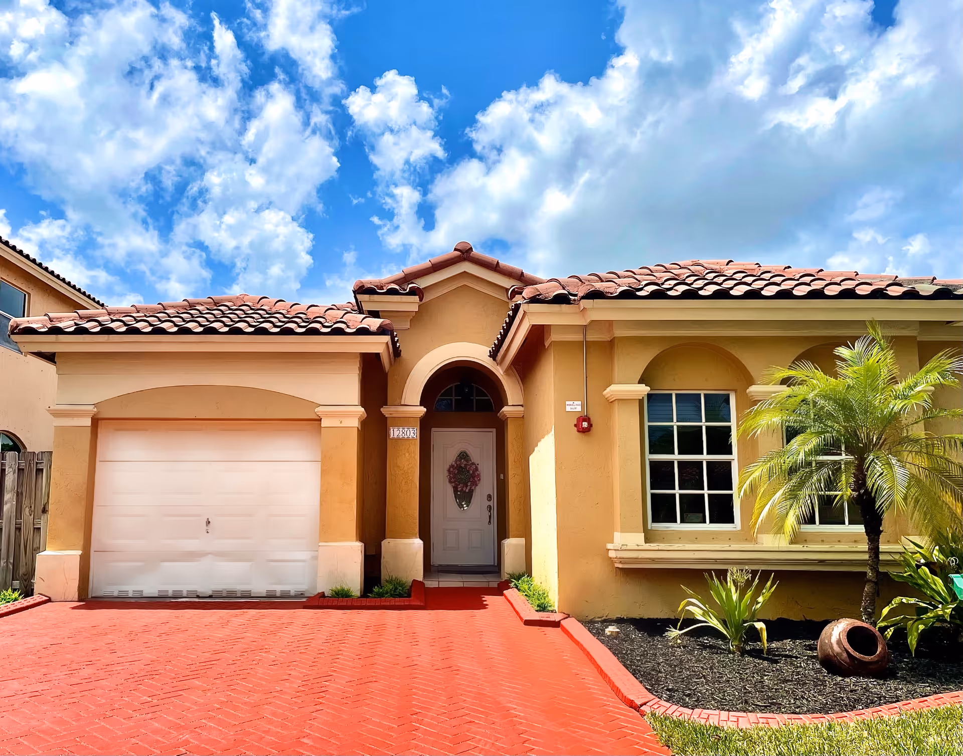 Front exterior view of a single-story house with a red brick driveway, a garage door on the left, a white front door with a floral wreath in the center, and a window with white trim on the right. There is a small landscaped area with a palm tree and other plants to the right of the driveway. The sky is blue with scattered clouds.