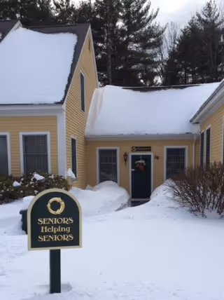 Snowy yellow building exterior with a front entrance and a sign reading Seniors Helping Seniors.