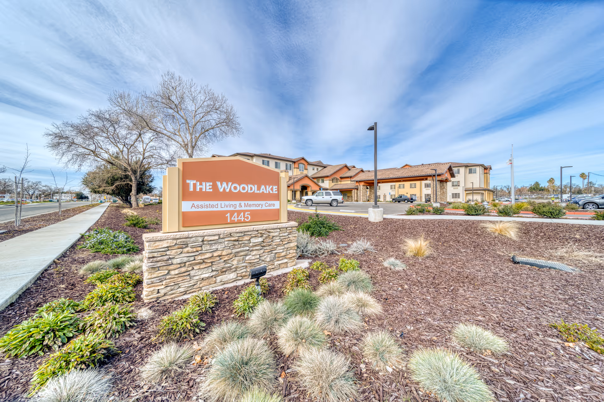 Exterior view of The Woodlake Senior Living facility with a large sign in the foreground that reads 'The Woodlake Assisted Living & Memory Care 1445'. The building is visible in the background with a parking lot and some cars. The landscaping includes various plants and mulch, and the sky is partly cloudy.