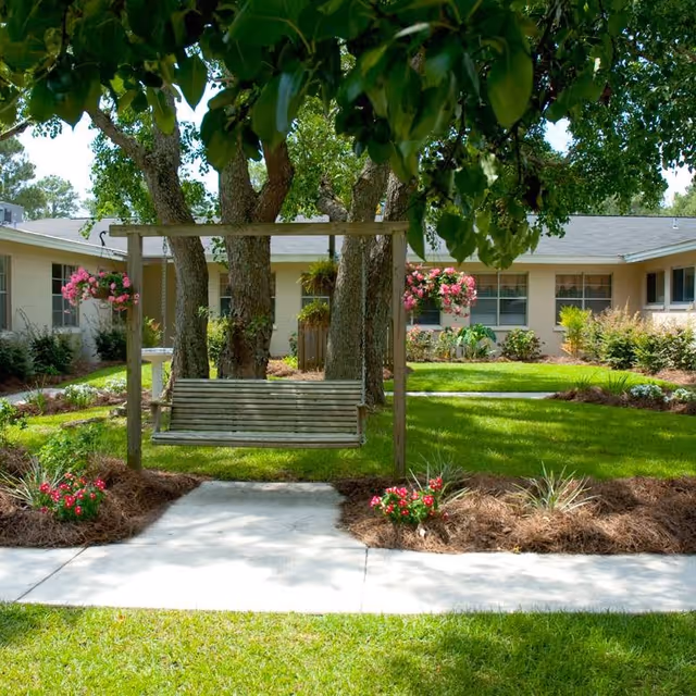 A wooden swing hanging from a wooden frame in a garden area with green grass, flower beds, and trees. In the background, there is a single-story building with windows and hanging flower baskets.