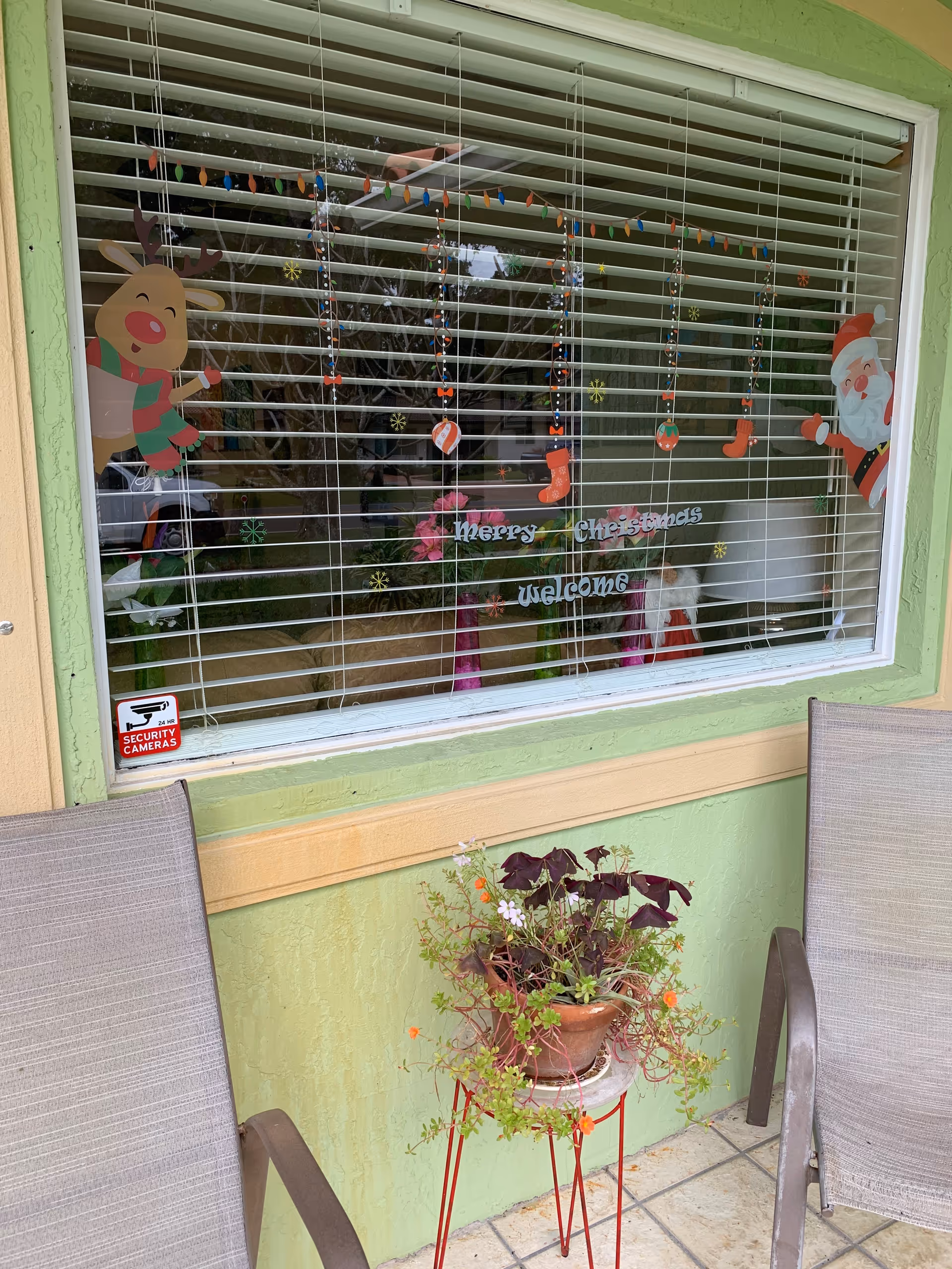 Window decorated with Christmas-themed stickers including a reindeer, Santa Claus, hanging ornaments, and the words 'Merry Christmas' and 'Welcome'. Two chairs are placed on either side of a small table with a potted plant in front of the window. The exterior wall is painted light green with a beige trim.