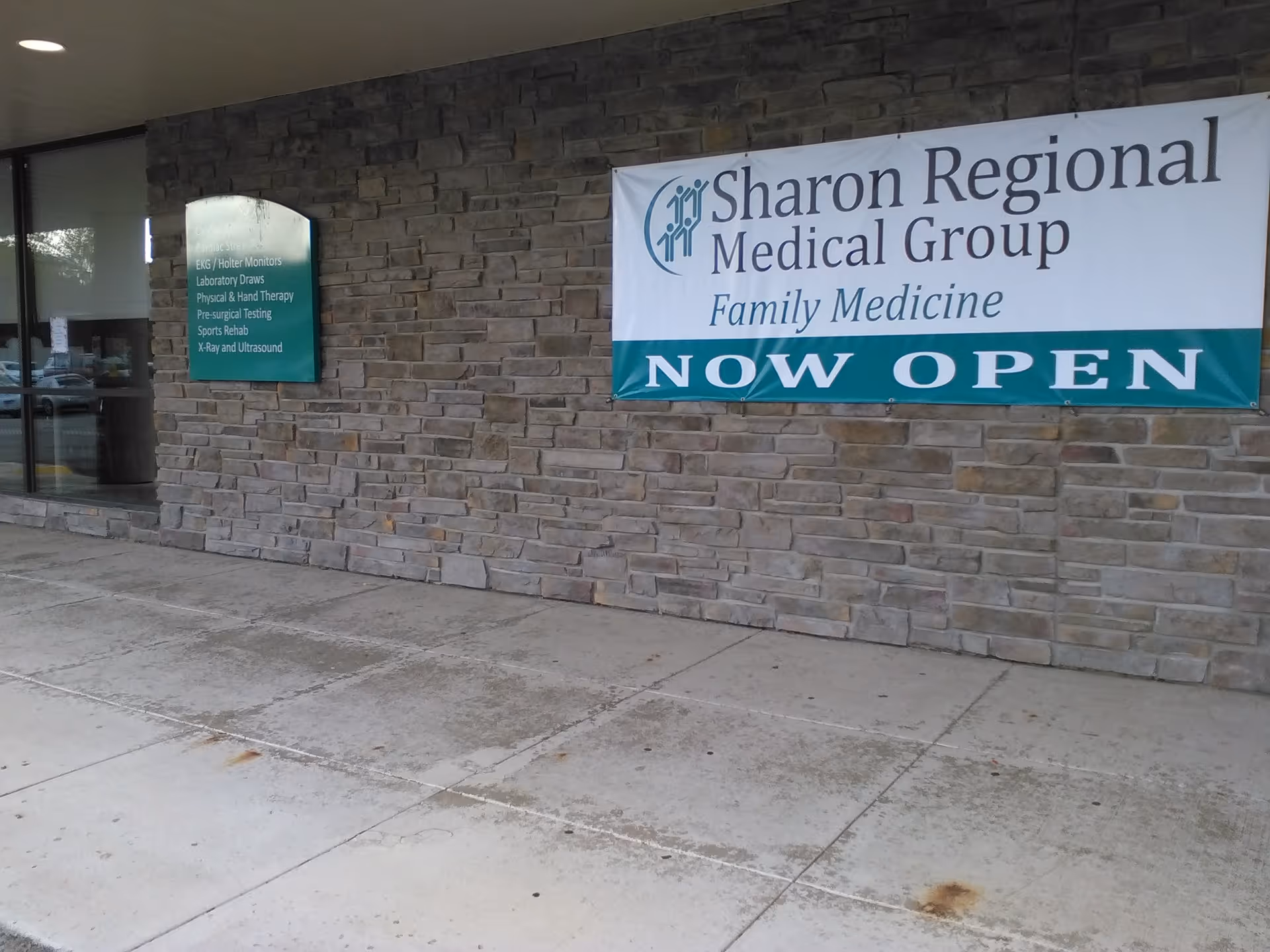 Stone exterior of a facility with a Sharon Regional banner reading NOW OPEN, sidewalk in front.