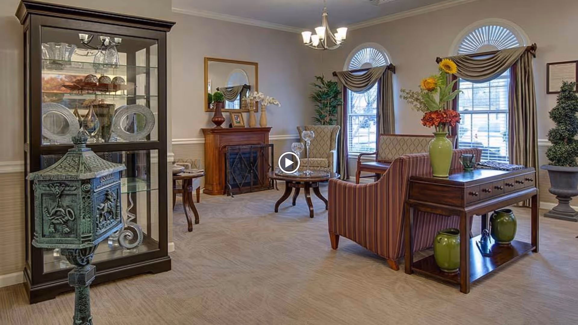 A cozy living room area in a senior living facility with a striped armchair, a wooden side table with green vases and a flower arrangement, a glass display cabinet with decorative plates and glassware, a small fireplace with a mirror above it, and large windows with arched tops and draped curtains letting in natural light.