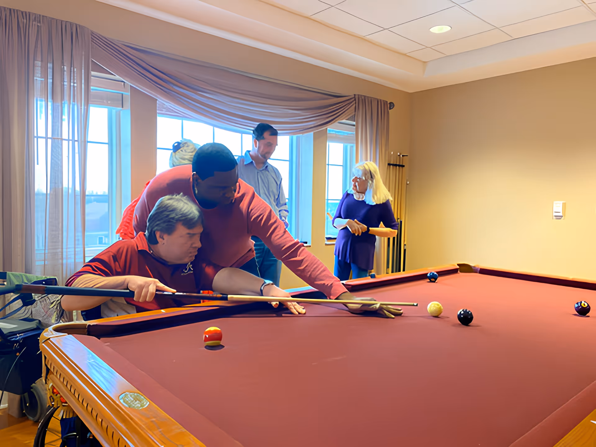 A group of people playing pool in a well-lit room with large windows covered by sheer curtains. One man is helping another man in a wheelchair aim his shot on the pool table, while two other people stand in the background holding pool cues.
