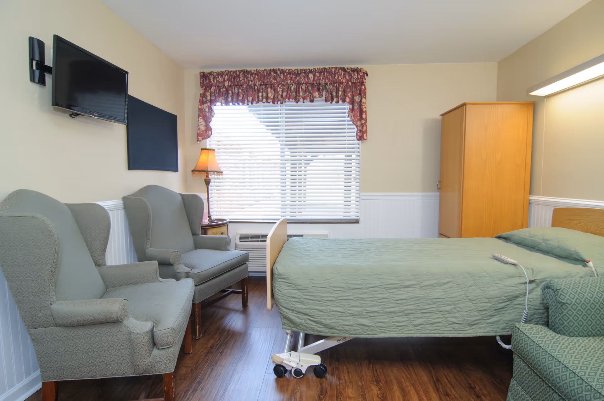 A senior living facility bedroom with a bed, two chairs, a wardrobe, lamp, and window.