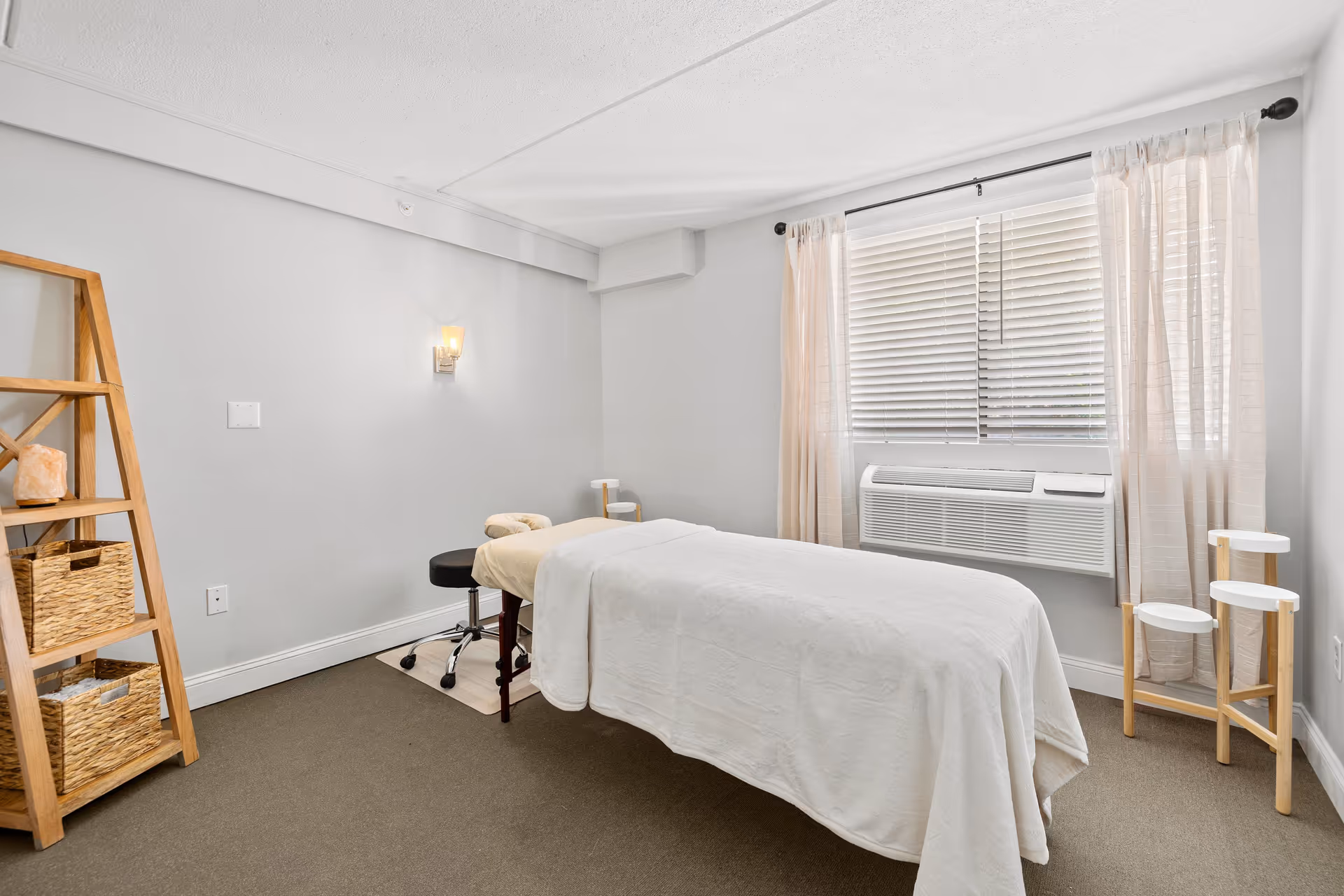 A bright, clean room with a massage table covered in a white sheet in the center. The room has light gray walls, a window with white blinds and sheer curtains, a wall-mounted air conditioning unit, a wooden ladder shelf with woven baskets on the left, and a small wooden stand with three circular white surfaces on the right.