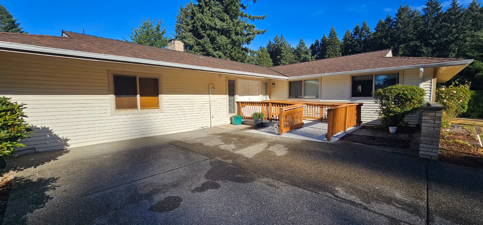 Front exterior of a single-story home with a wooden entry ramp, paved driveway, and surrounding trees.