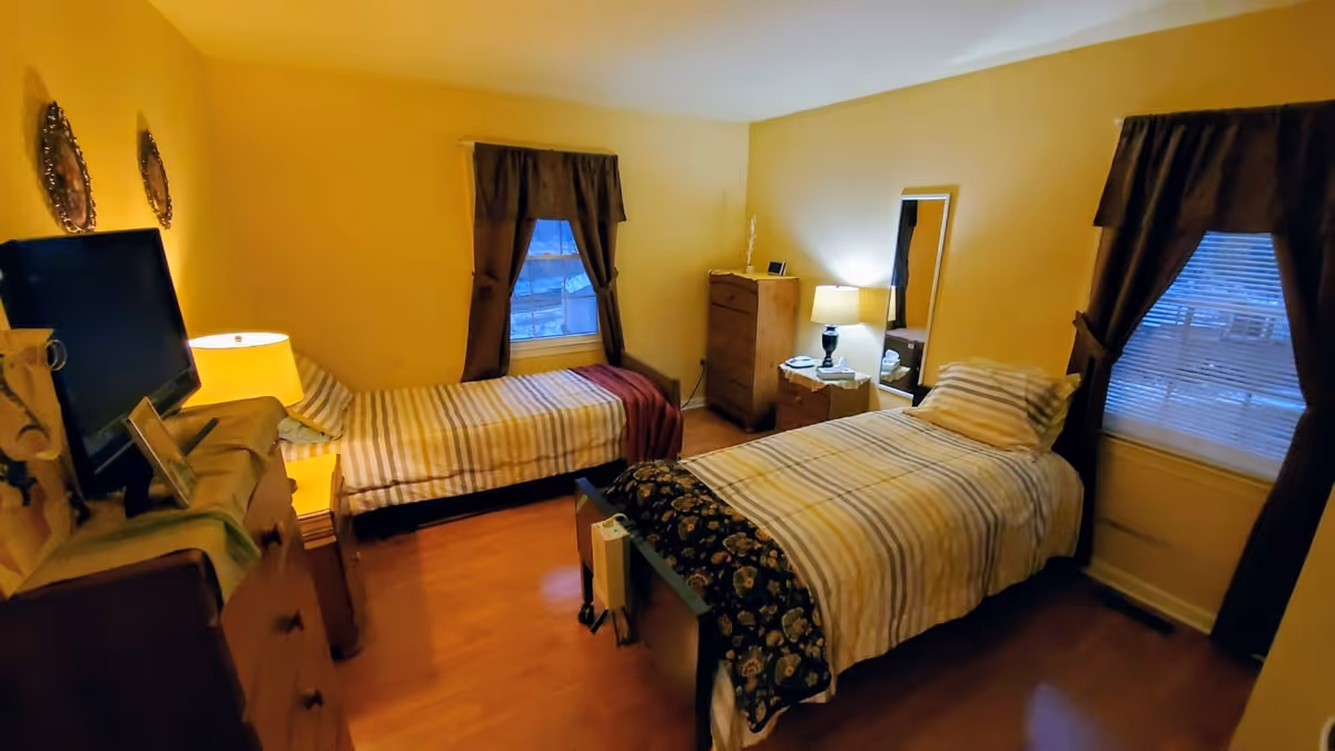 Two single beds with striped bedding in a yellow-toned bedroom with lamps, dresser, and windows.