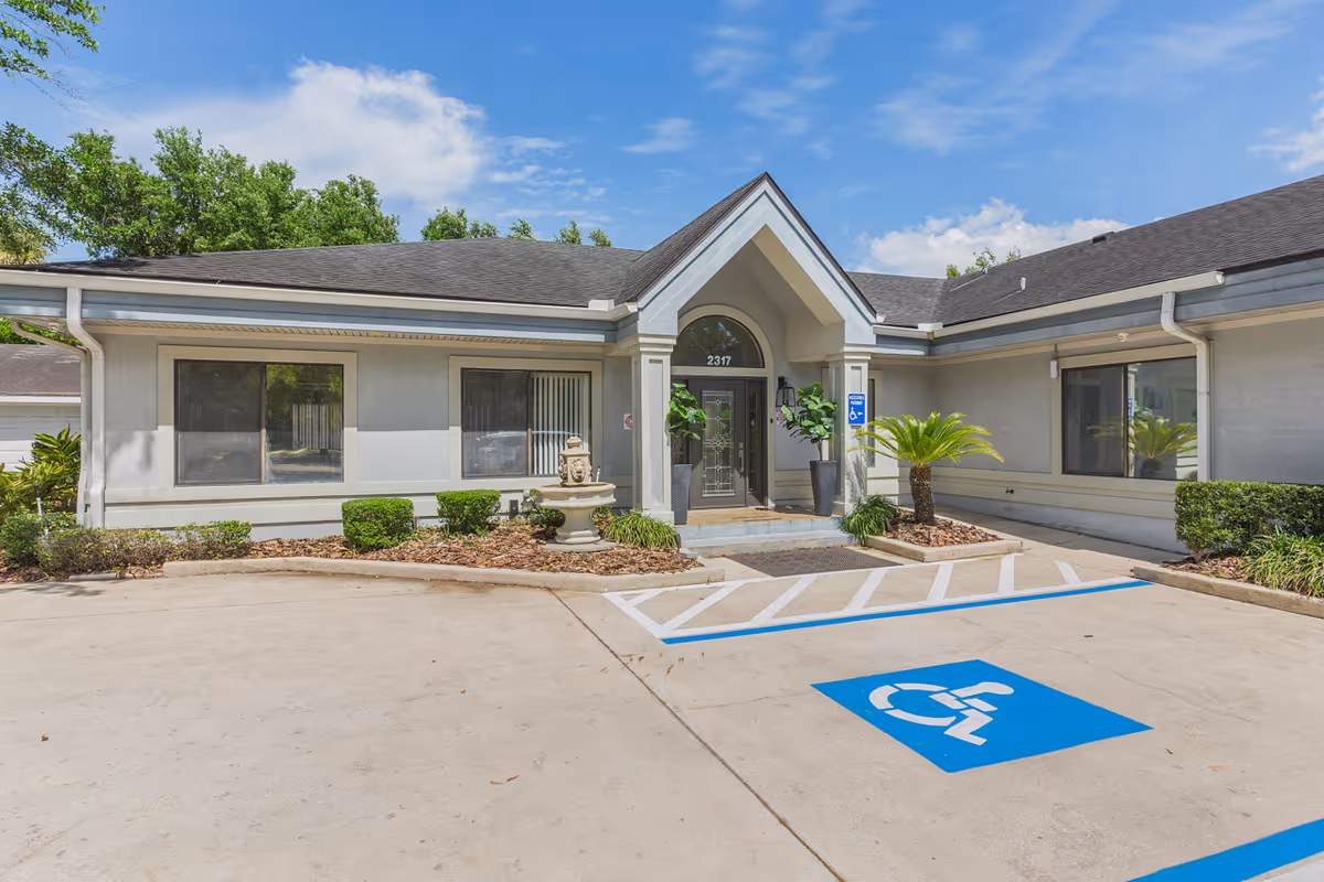 Exterior view of a single-story building with a peaked entrance, two large windows on either side, and a handicapped parking space in front. The building is surrounded by small shrubs and plants under a partly cloudy blue sky.