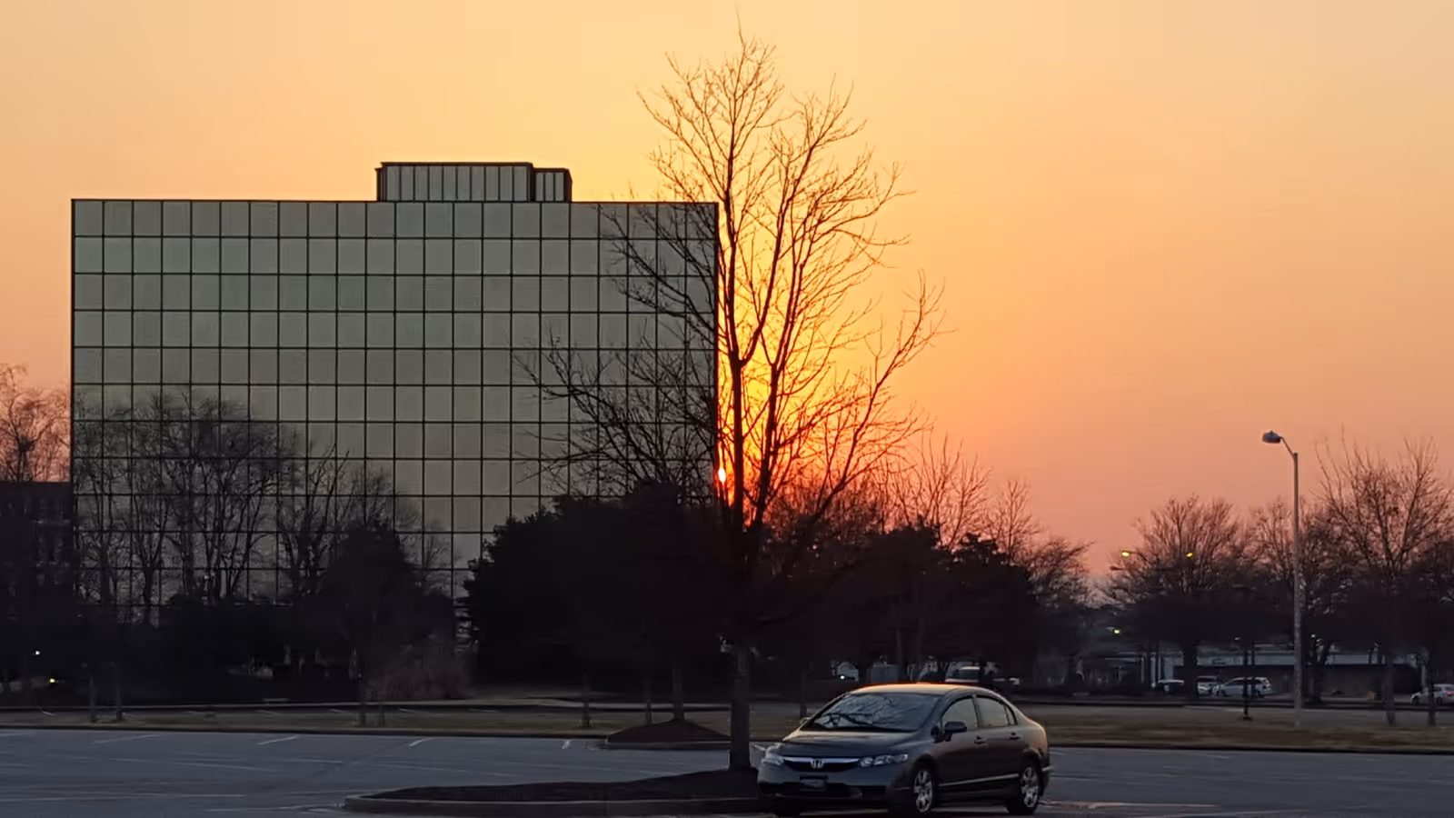 Sunset over a glass-front senior living building with bare trees and parked cars.