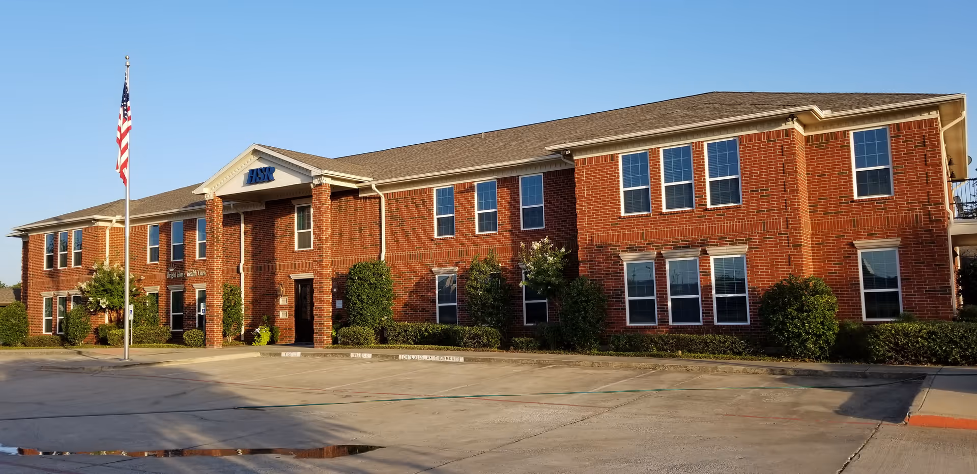 Brick two-story senior living facility with entrance, flagpole, and manicured shrubs.