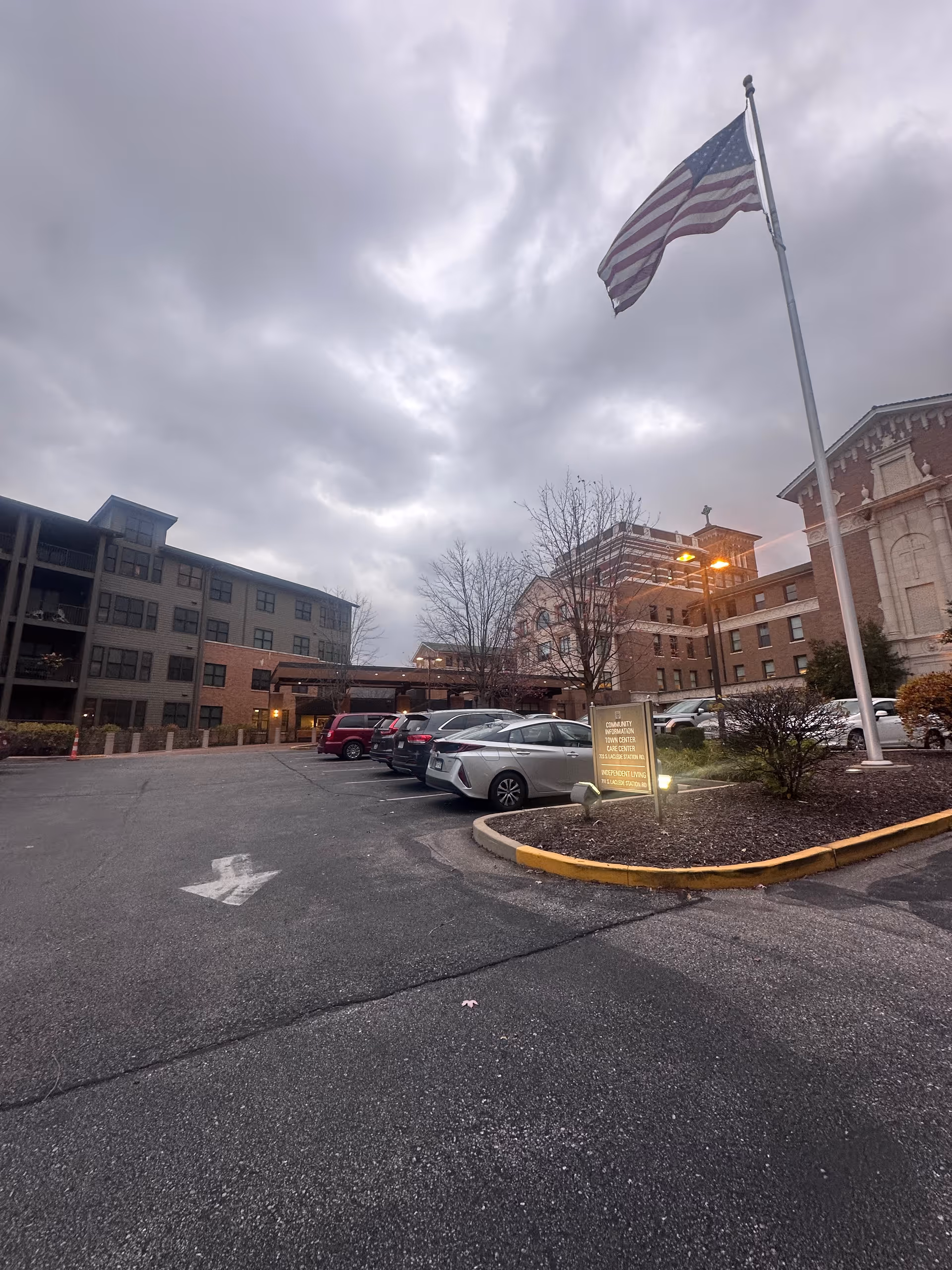 Parking lot and entrance of the Lutheran Convalescent Home with parked cars, a flagpole flying an American flag, and multi-story buildings under a cloudy sky.