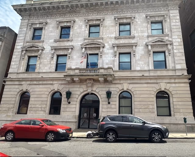 Stone-front building with arched entrance, flag, and two parked cars along curb.