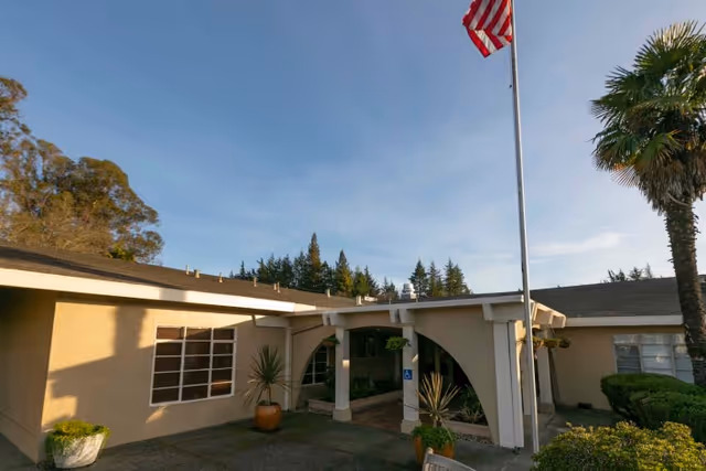 Front entrance of a single-story building with an arched covered walkway, potted plants, and a flagpole flying the U.S. flag under a clear sky.
