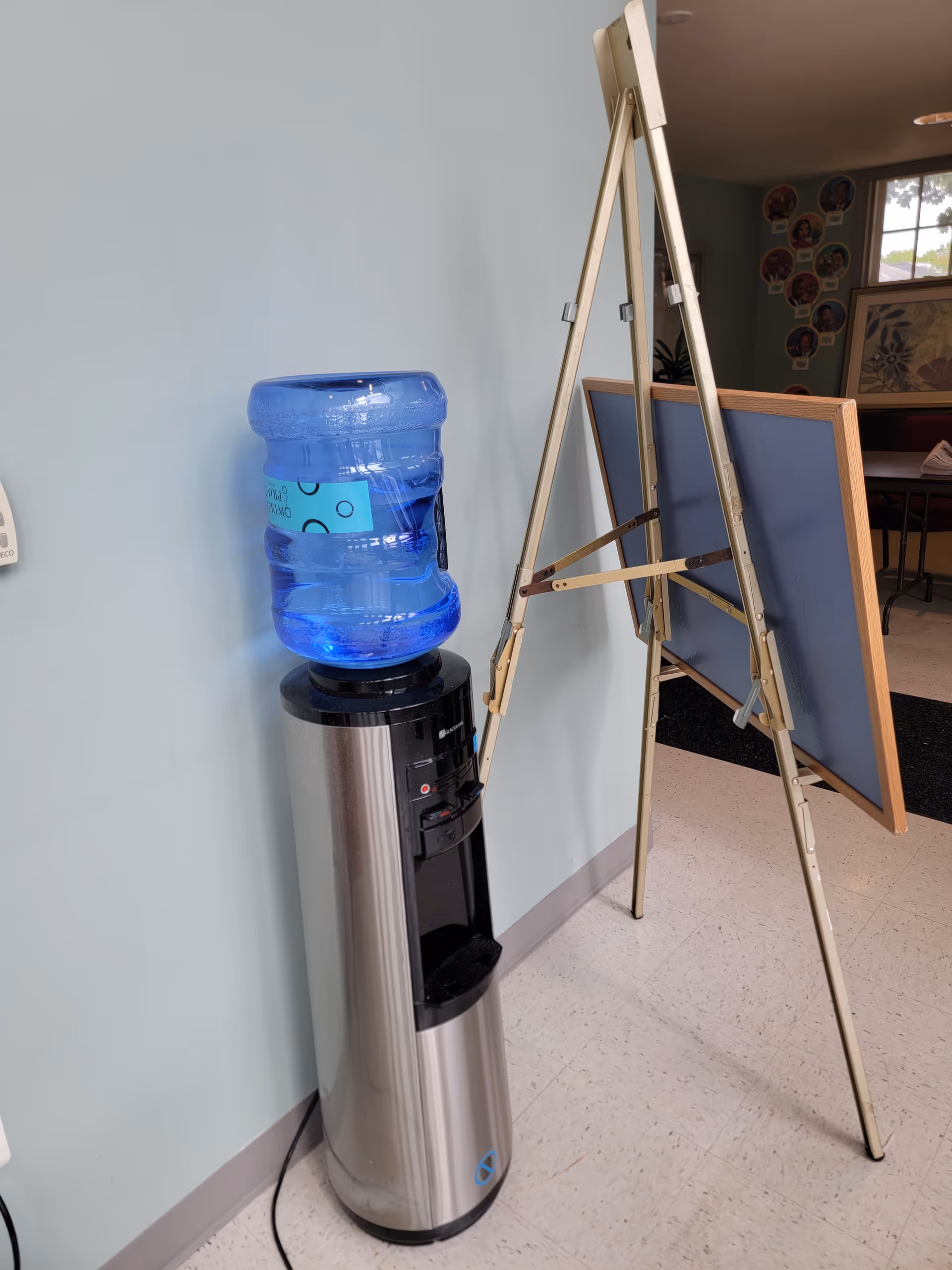 Water cooler with blue jug beside folding easel in a light blue hallway