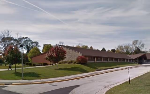 Low single-story building with a sloped roof, grassy lawn, trees, and a curved driveway under a partly cloudy sky.