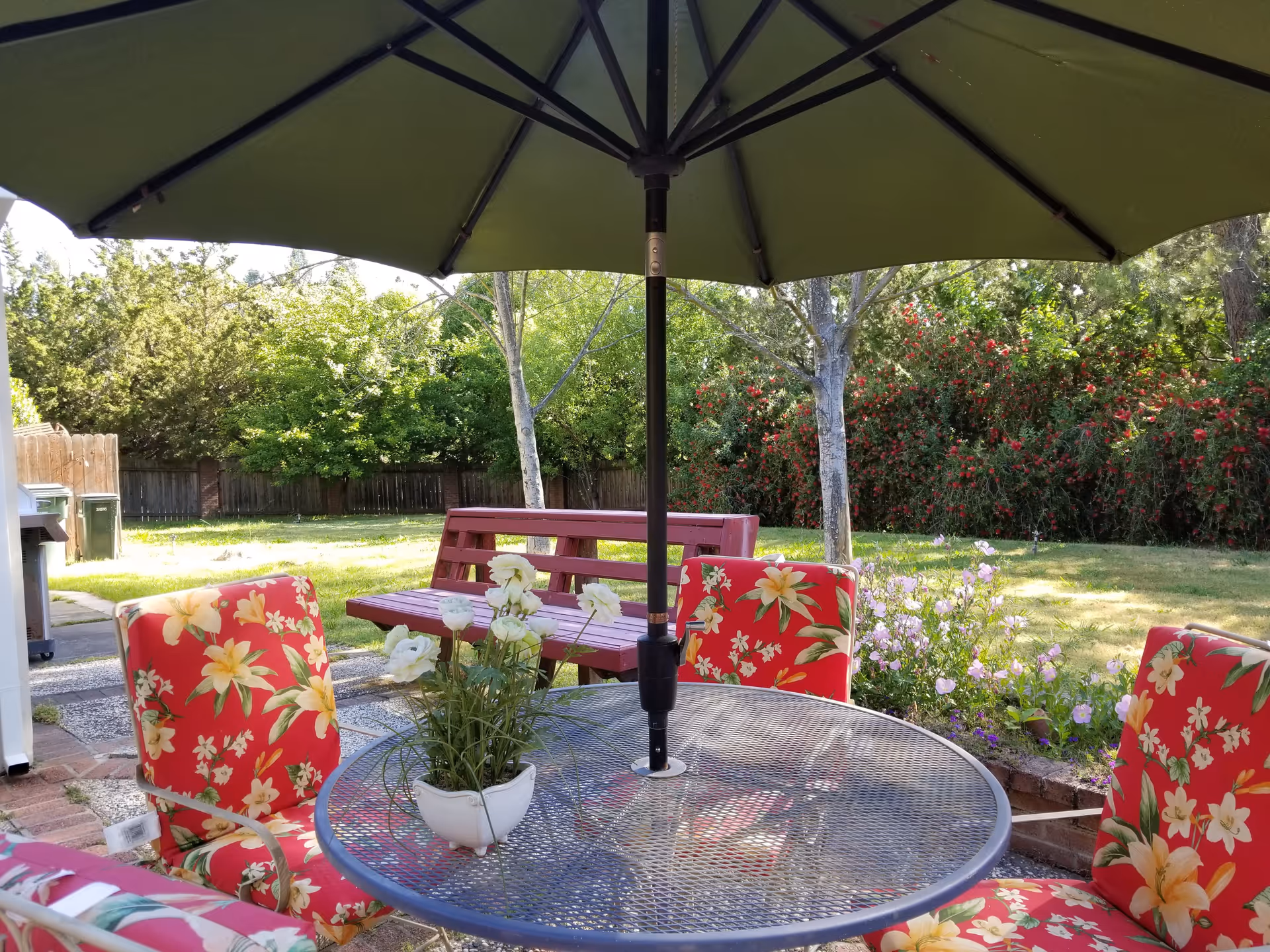 Outdoor patio with a round metal table, red floral chairs, umbrella, and garden.