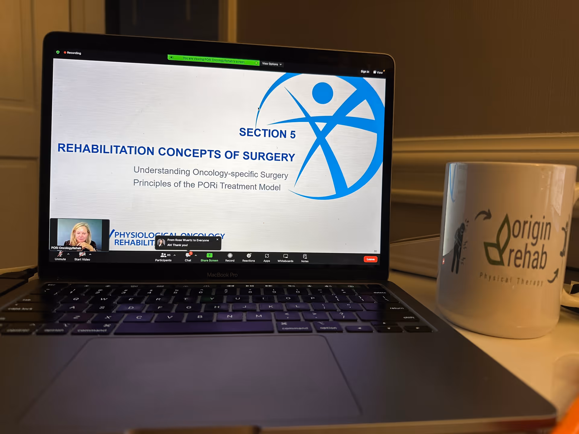 Laptop on a desk shows a rehabilitation presentation; an Origin Rehab mug sits beside it.