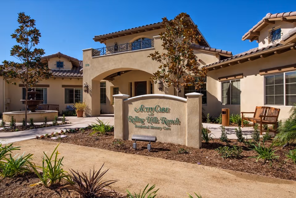 Exterior view of ActivCare Rolling Hills Ranch, a beige stucco building with a tiled roof, surrounded by landscaped plants and trees. There is a sign in front displaying the facility's name and a bench nearby under a clear blue sky.