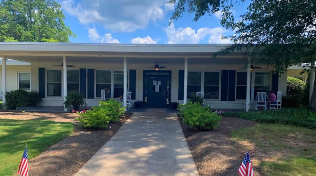 Front exterior view of a single-story building with a covered porch, white columns, and black shutters on the windows. There are rocking chairs on the porch and small American flags planted in the garden beds along the walkway leading to the entrance. The sky is blue with some clouds and there are trees surrounding the building.