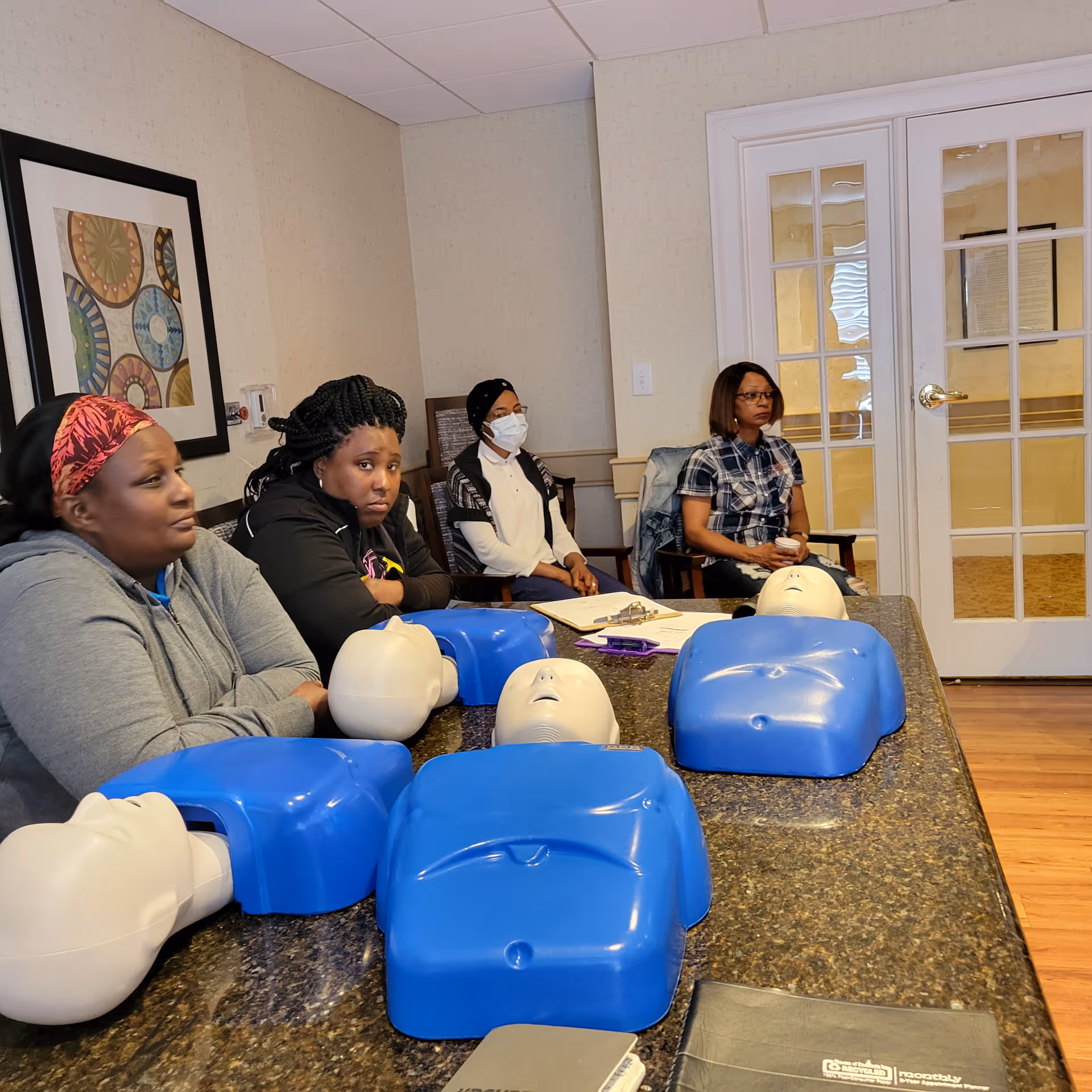 Several adults sit in a CPR training room with blue manikins arranged on a counter.