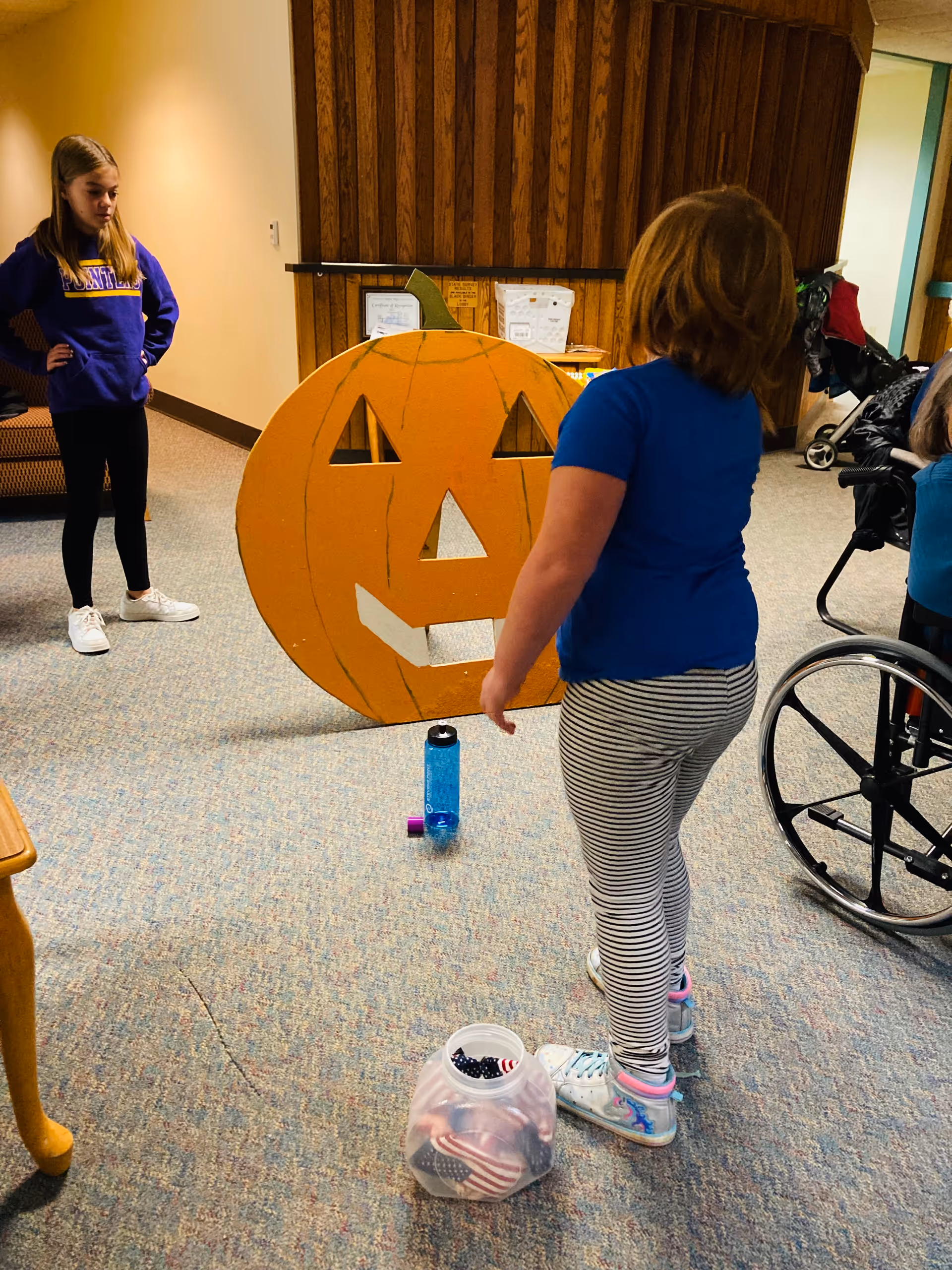 Indoor community area with a large jack-o'-lantern pumpkin and two girls nearby.