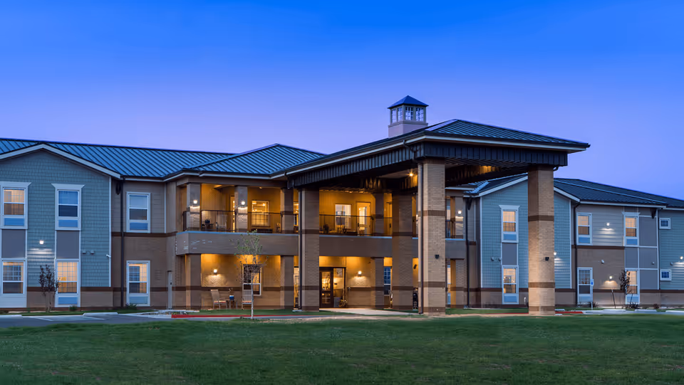 Front exterior of a two-story senior living building at dusk with an illuminated entry canopy and a grassy lawn.