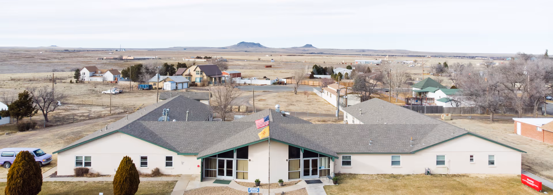 Aerial view of Clayton Nursing & Rehab facility showing a single-story building with a central entrance, an American flag and another flag flying in front, surrounded by a dry landscape with scattered trees and houses in the background.