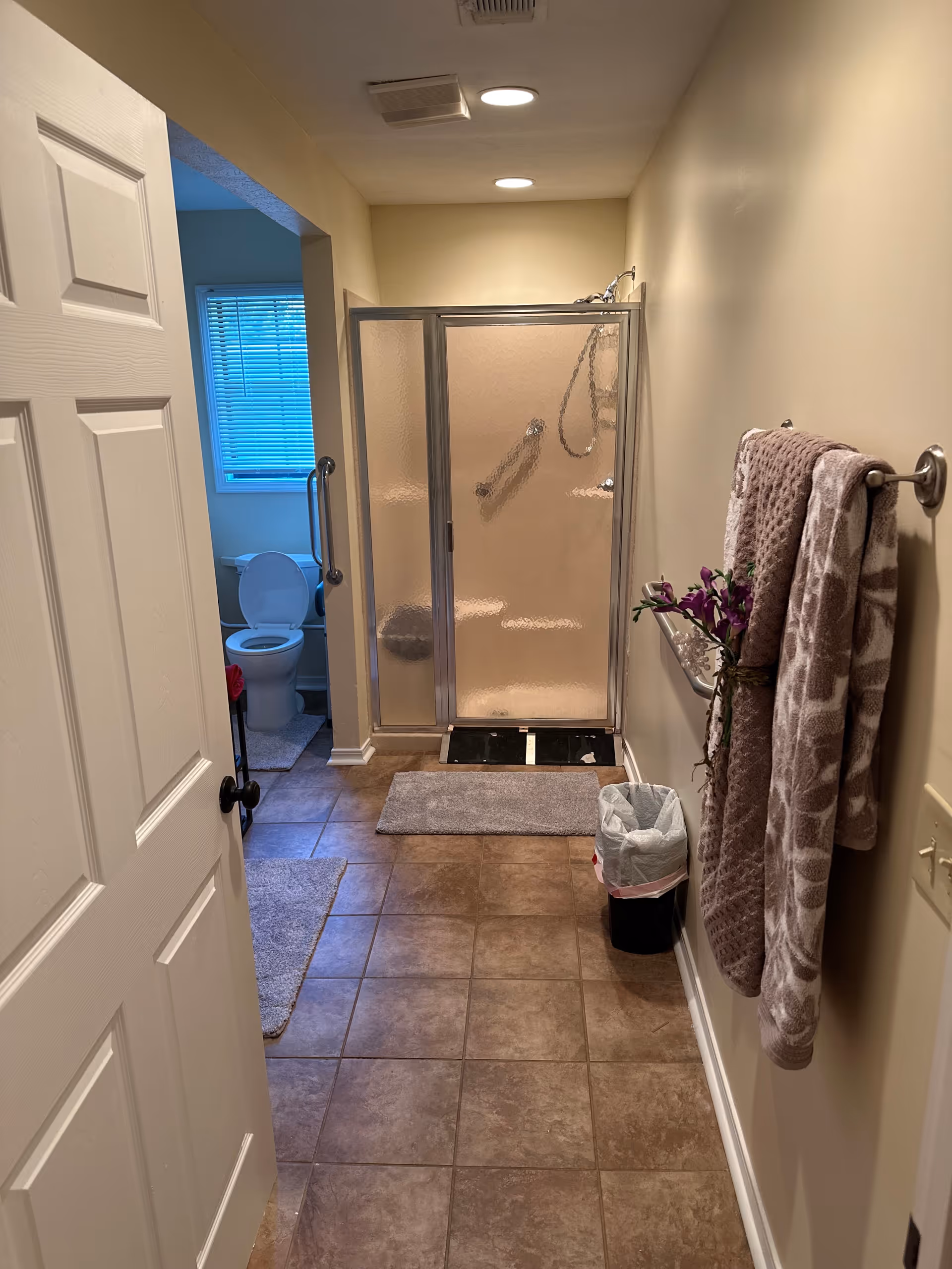 Narrow bathroom hallway with frosted glass shower enclosure, towels, and a trash can.