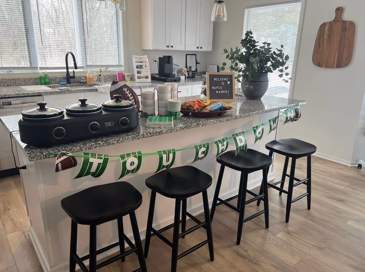 Kitchen island with black stools, banner, appliances, and a plant in Maple Manor's kitchen.
