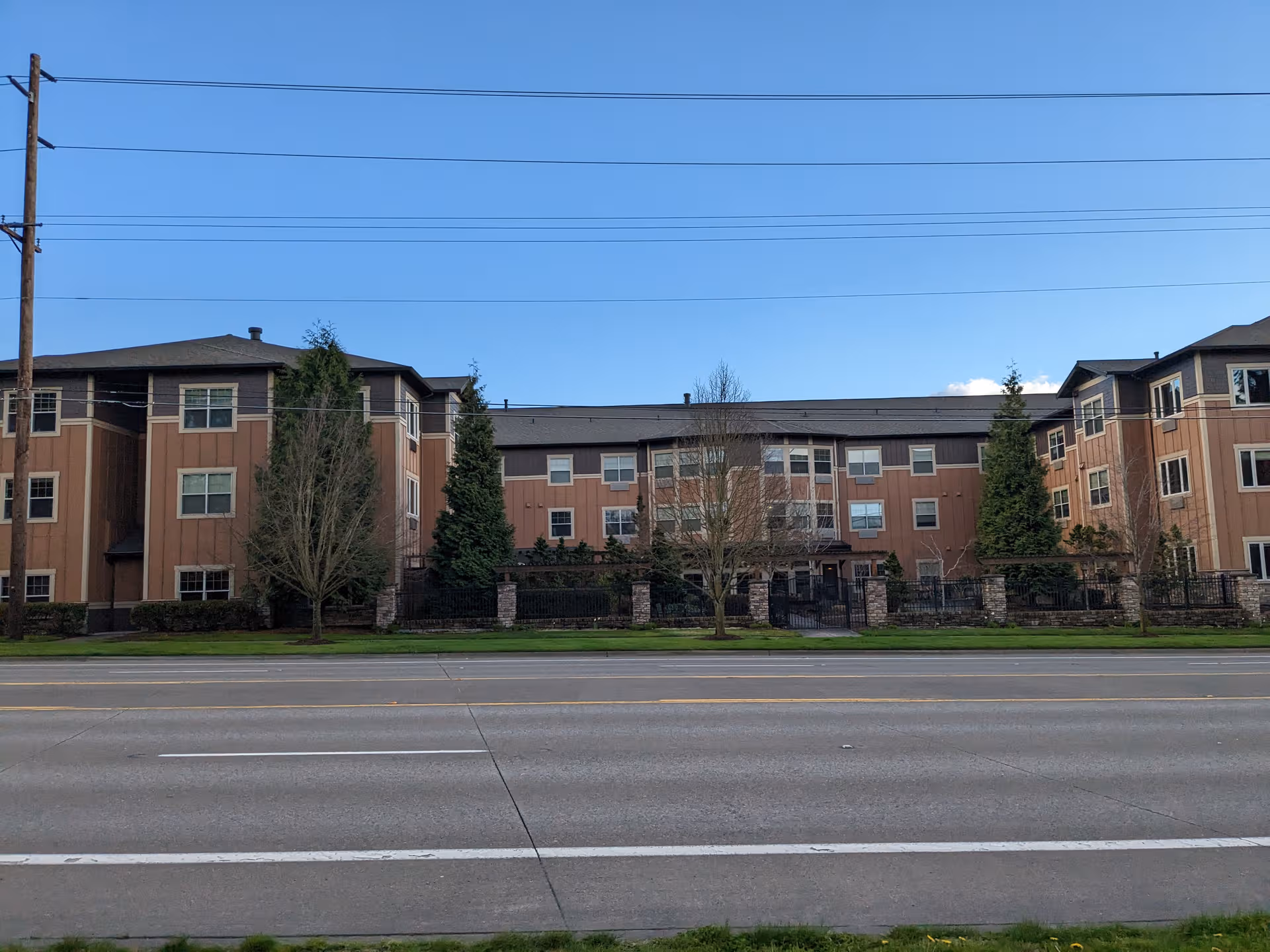 Three-story senior living building with tan siding, trees, and a fence along the street.