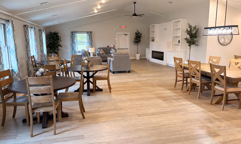 Bright senior living common area with round tables, wooden chairs, sofa seating, and a fireplace.