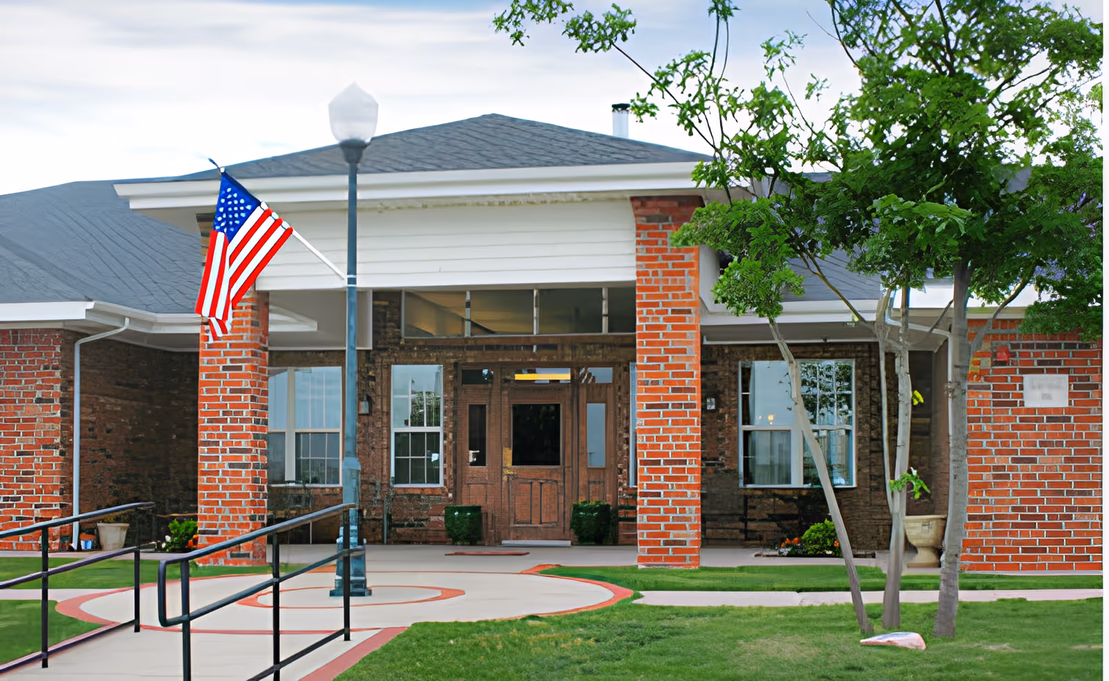 Front entrance of a brick building with a covered porch supported by brick columns. An American flag is mounted on one of the columns. There is a lamp post near the entrance and a tree with green leaves on the right side. The walkway leading to the entrance has black handrails and is surrounded by green grass.