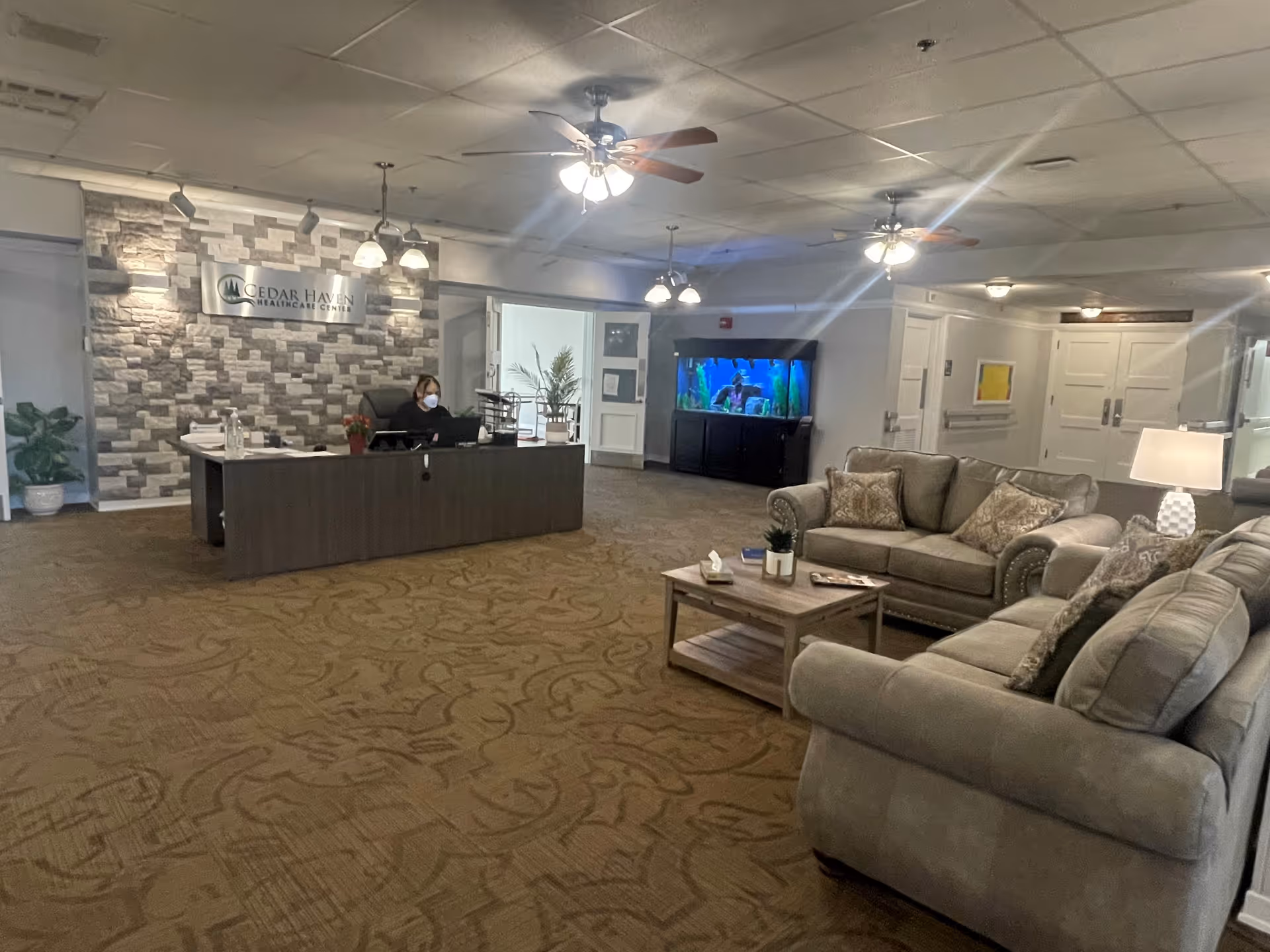 Spacious lobby of Cedar Haven Healthcare Center with a reception desk and staff member, a seating area with sofas and coffee table, and a large aquarium.