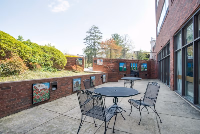 Brick-walled outdoor patio with metal tables and chairs, decorative tile murals along a low wall, and large building windows.