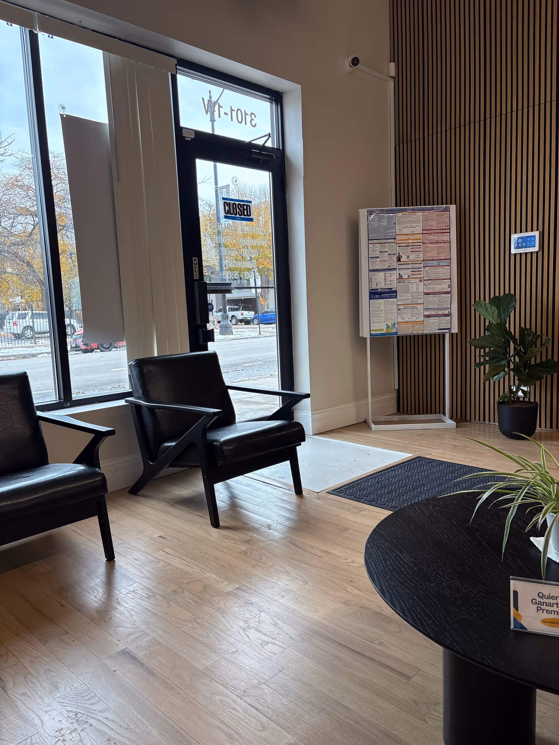 Lobby area with black chairs, glass door, bulletin board, and potted plant.